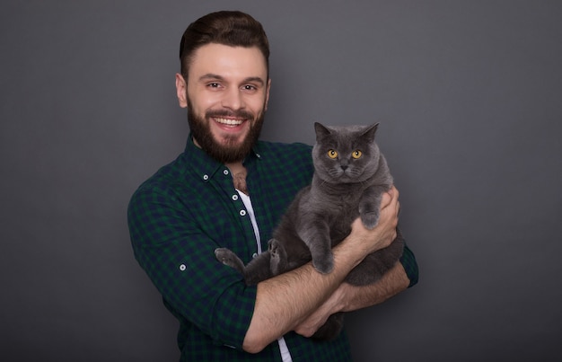 Bello giovane uomo barbuto sorridente tiene il suo adorabile gatto soffice sulle mani e posa insieme come migliori amici