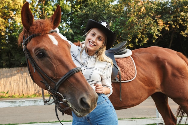 Bella giovane donna bionda sorridente che accarezza un cavallo al cortile dei cavalli