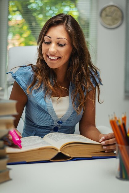 La bella giovane studentessa sorridente con il libro sta preparando l'esame e imparando le lezioni nella biblioteca della scuola.