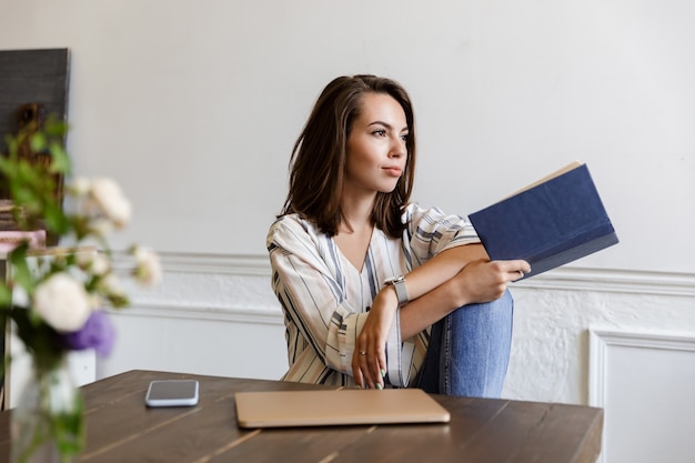 Beautiful smiling young girl sitting at the table at home, reading a book, relaxing