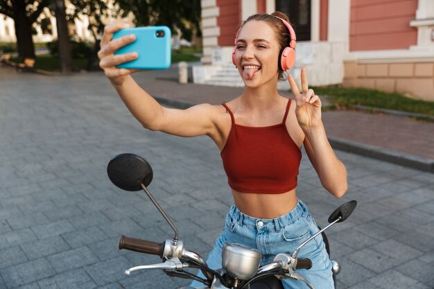 Beautiful smiling young girl wearing casual summer clothing sitting on a scooter outdoors at the city streets, taking a selfie while listening to music with wireless headphones