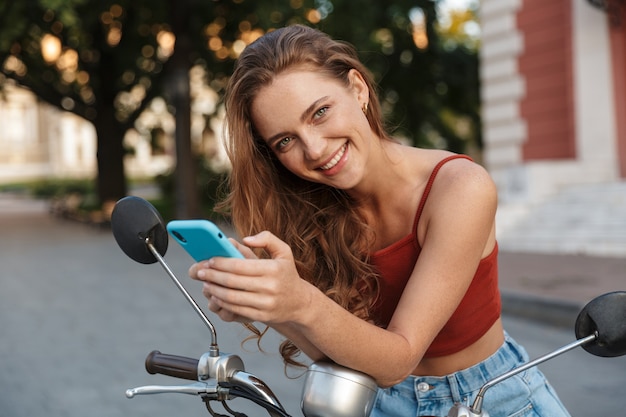 Beautiful smiling young girl wearing casual summer clothing sitting on a scooter outdoors at the city streets, using mobile phone