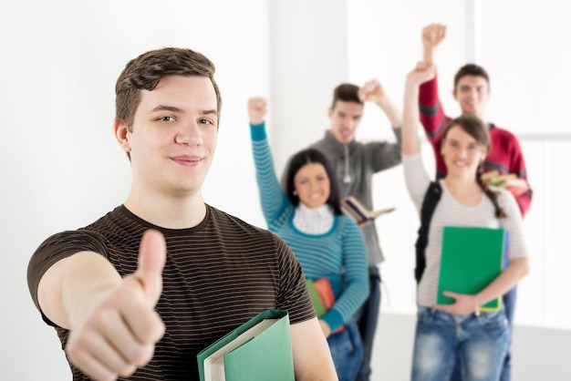 A beautiful smiling young man with book and thumb up is standing in the foreground. A happy group of his friends is behind him with arms in the air.