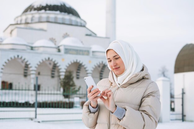 Beautiful smiling young Muslim woman in headscarf in light clothing using mobile against mosque