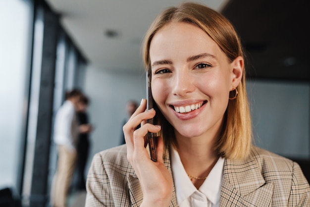 Bella giovane donna d'affari intelligente sorridente in piedi in ufficio con un gruppo di colleghi sul muro, parlando al cellulare