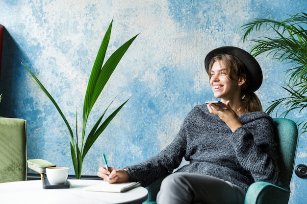 Beautiful smiling young woman dressed in sweater and hat sitting in chair at the cafe table, talking on mobile phone, stylish interior, taking notes