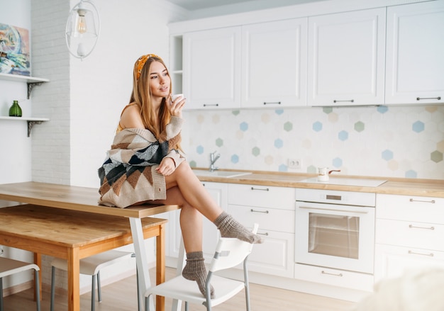 Красивая улыбающаяся девушка с длинными светлыми волосами в уютном вязаном кардигане с чашкой утреннего кофе сидит на кухонном столе дома