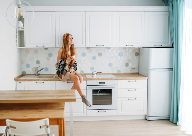 Beautiful smiling young woman fair long hair girl wearing in cozy knitted cardigan with cup of morning cofee sitting on kitchen work surface at home