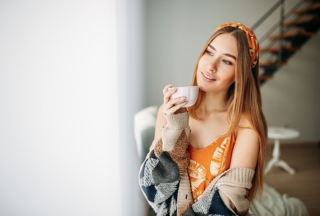 自宅の窓の近くのお茶と居心地の良いニットカーディガンを着て美しい笑顔の若い女性公正な長い髪の少女