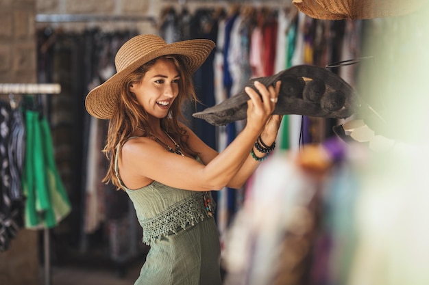 A beautiful smiling young woman is walking along the streets of a Mediterranean town and enjoys in summer sunny day.  She chooses a new hat, wanting to buy it.