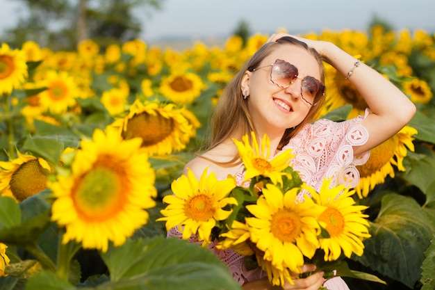 Beautiful smiling young woman in a white shirt stands in the field among sunflowers