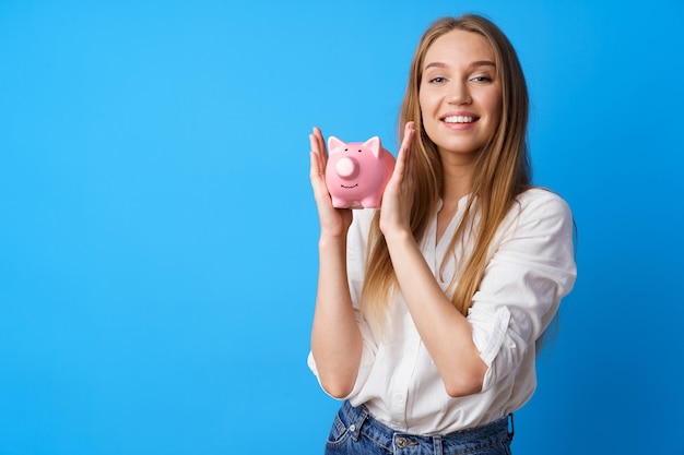 Beautiful smiling young woman with piggy bank on blue background