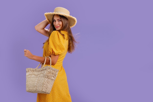 Beautiful smiling young woman in yellow dress summer straw hat bag isolated on pastel violet background