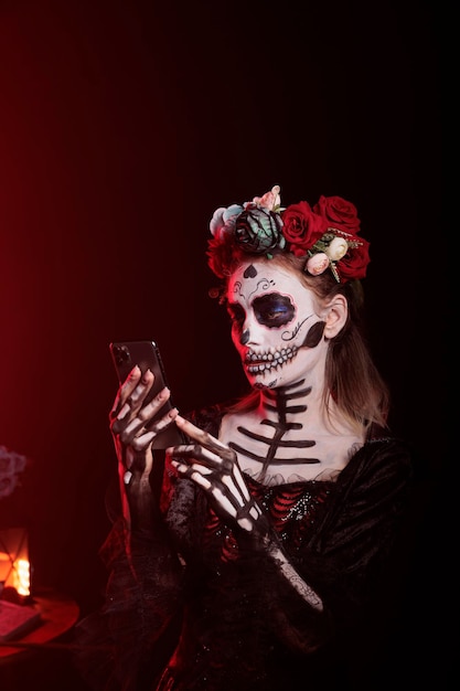 검은 배경 위에 스마트폰을 사용하여 스튜디오에서 인터넷 웹사이트를 탐색하는 아름다운 으스스한 여성. 멕시코 휴일 축하에 죽음의 공포 여신을 묘사한 산타 무에르테 모델.