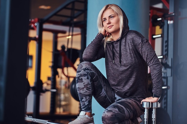 Beautiful sportive girl with blonde hair wearing hoodie sitting with dumbbells, looking sideways with a thoughtful look in the gym