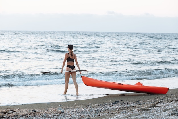 黒の水着を着た美しいスポーツの女の子は、日没時に海で泳ぐためにオレンジ色のカヤックと一緒に行きます