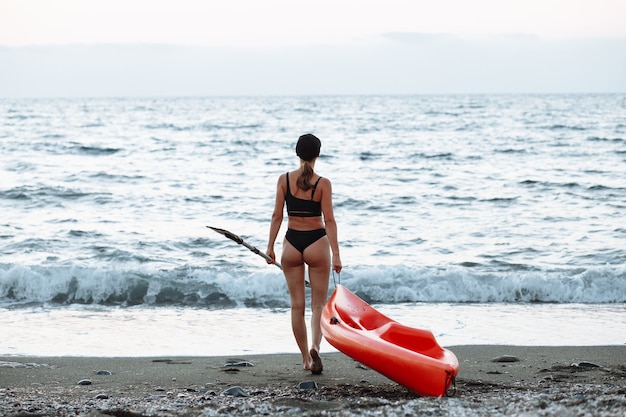 Foto la bella ragazza sportiva in costume da bagno nero va con un kayak arancione per nuotare nel mare al tramonto