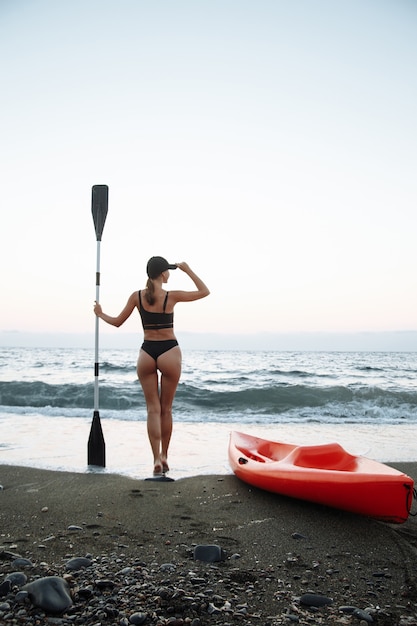 写真 黒の水着を着た美しいスポーツの女の子は、日没時に海で泳ぐためにオレンジ色のカヤックと一緒に行きます