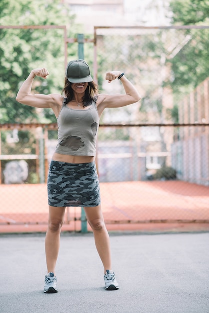 Beautiful sports girl in khaki clothes and cap on the playground shows biceps and smiles. Healthy lifestyle. 