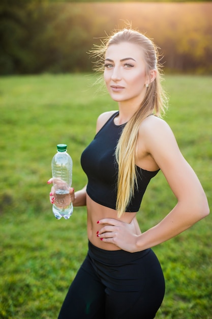 Красивая спортивная женщина в топе и кроссовках на утренней пробежке пьет воду из бутылки