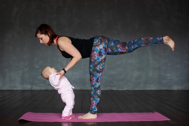 美しいスポーティなフィットの女性は彼女の赤ちゃんを保持しているヨガのアーサナ Virabhadrasana を実践します