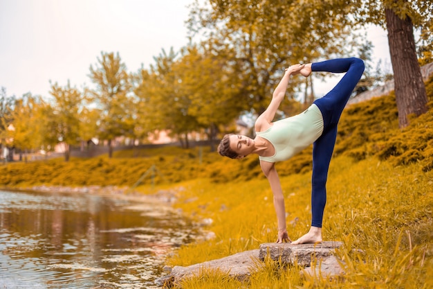 Bella ragazza sportiva che sta nella posa della mezza luna, esercizio di ardha chandrasana snello. esercizi di yoga sull'erba verde in autunno sullo sfondo della natura