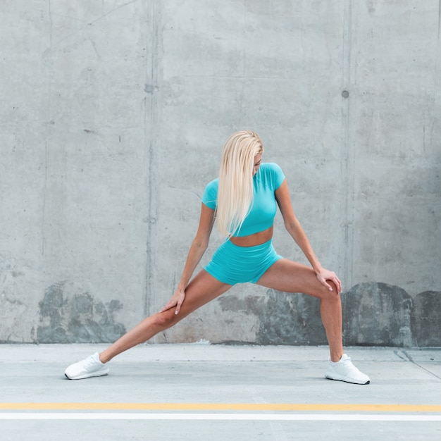 Красивая спортивная молодая женщина фитнес-модель со светлыми волосами в модной синей спортивной одежде с белыми кроссовками начинает упражнения на растяжку возле серой бетонной стены в городе