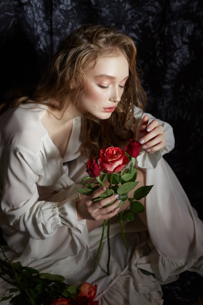 사진 아름 다운 봄 소녀는 그녀의 손에 장미 꽃과 함께 바닥에 앉아있다. 하얀 드레스를 입고 여자는 낭만적 인 이미지를 꿈꾸고 있습니다. 곱슬 머리를 가진 금발