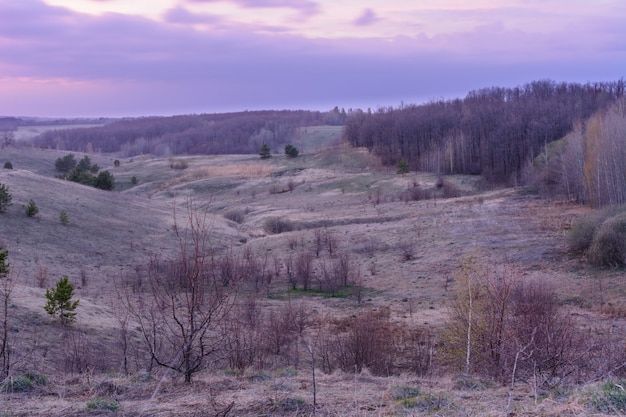 Красивый весенний пейзаж: закат, деревья, лес, горы, холмы, поля, луга и небо. Великолепное красное небо с тяжелыми облаками на закате. Ранняя весна.