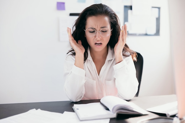 Beautiful stressed young office worker sitting at desk holding head because of pain in office