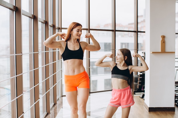 La bella donna forte e la piccola figlia affascinante stanno mostrando il loro bicipite e stanno sorridendo mentre si allenano in palestra