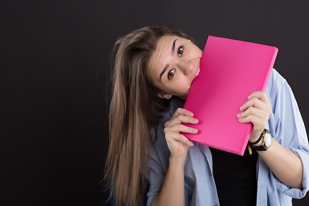 Beautiful student in denim shirt with long hair, on black wall, gnawing book