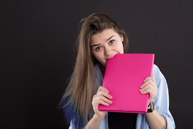 Beautiful student in denim shirt with long hair, on black wall, gnawing book