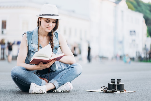 Beautiful student girl reading a book while sitting on the asphalt in the middle of the street