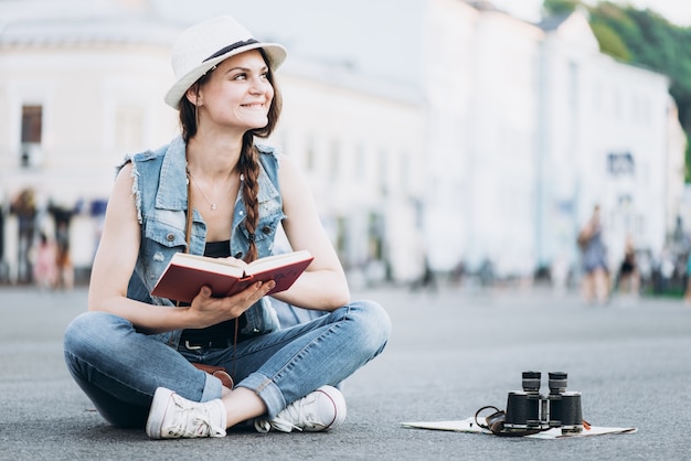 Photo beautiful student girl reading a book while sitting on the asphalt in the middle of the street