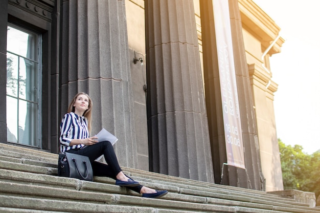 Красивая студентка сидит на ступеньках возле колледжа с рюкзаком, держит бумаги в руках и смотрит вверх, она усваивает уроки на перемене
