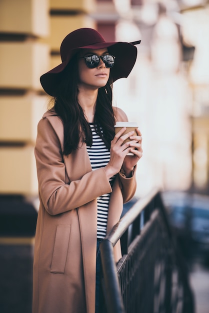 Beautiful and stylish. Beautiful young woman in sunglasses holding coffee cup and looking away while standing outdoors