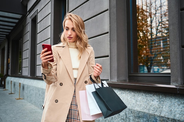 Beautiful stylish blond girl in beige coat with shopping bags using cellphone on city street