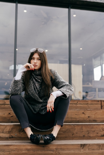 Beautiful stylish brunette girl in gray coat sits on steps outdoors, posing