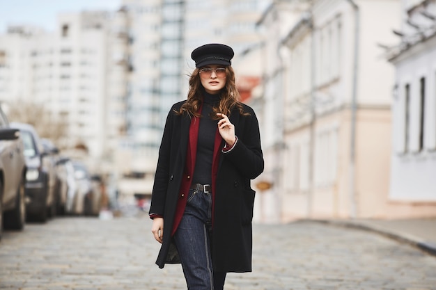 Beautiful and stylish girl, in cap, coat and sunglasses, walking by the street.