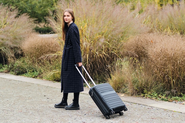 Beautiful stylish girl in coat with suitcase dreamily looking in camera outdoor