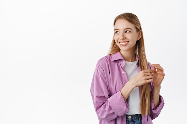 Beautiful stylish girl playing with hair, turn head and look behind shoulder with happy smile, reading promotional text on white blank copyspace, standing against studio wall