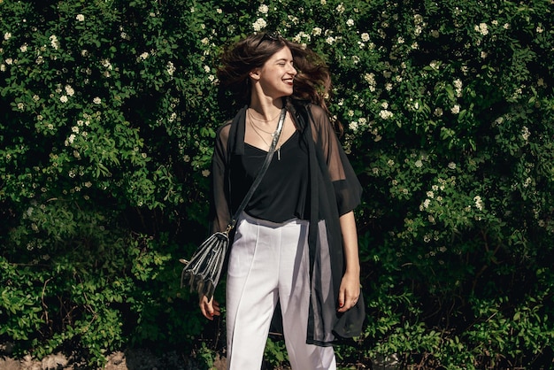 Beautiful Stylish Hipster Girl with beautiful hair having fun at floral bushes in sunny day Boho Woman with sunglasses in fashionable outfit smiling and enjoying day in garden