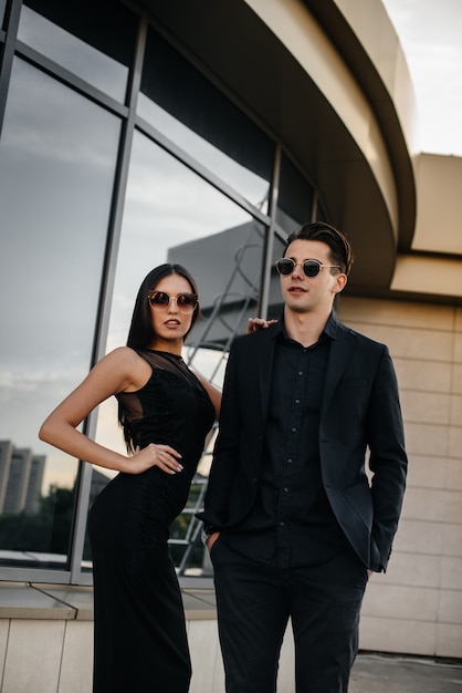 Photo a beautiful, stylish pair of young people in black clothes and glasses stand against the background of an office building in the sunset. fashion and style