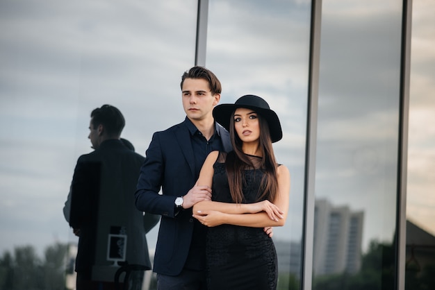 A beautiful, stylish pair of young people in black clothes and glasses stand against the background of an office building in the sunset. Fashion and style