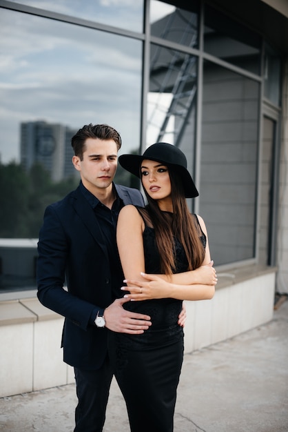 A beautiful, stylish pair of young people in black clothes and glasses stand against an office building in the sunset. Fashion and style