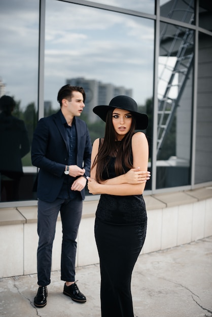 A beautiful, stylish pair of young people in black clothes and glasses stand next to an office building in the sunset. Fashion and style.