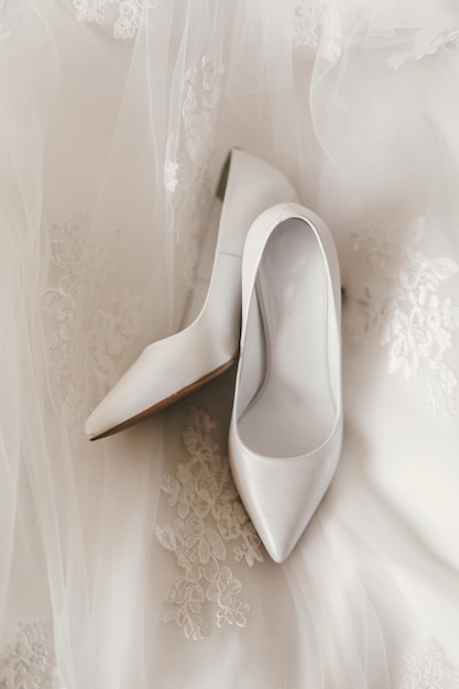 Beautiful stylish shoes on luxury silk wedding dress simple white shoes on white gown with lace floral ornaments bridal morning preparations