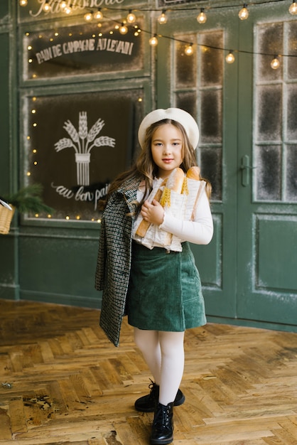 A beautiful stylish six year old girl holds a bag of baguettes in her hands and stands near the window of a bakery with fresh pastries