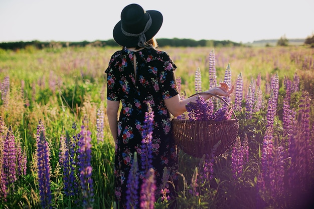 Photo beautiful stylish woman gathering lupine in wicker rustic basket in sunny field tranquil moment